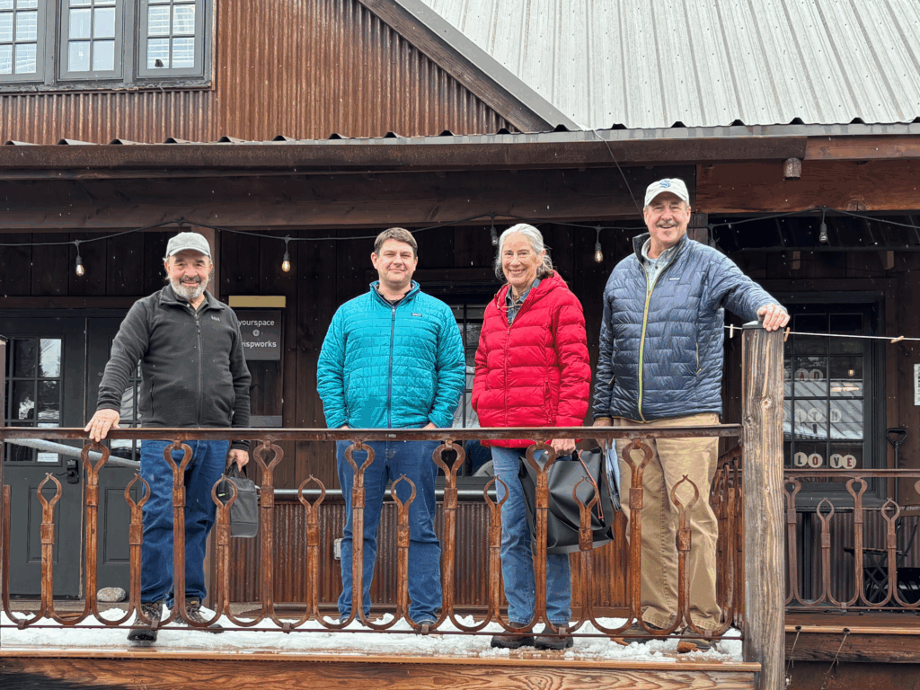 Four people wearing jackets stand on a wooden porch railing in front of a rustic building.