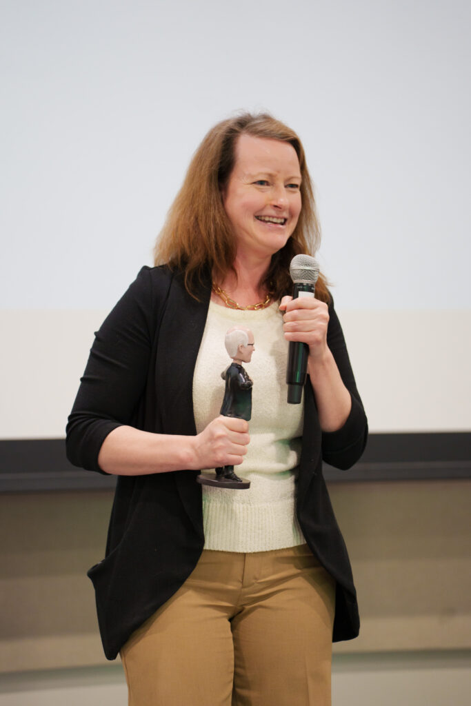 A woman wearing a black cardigan and light sweater smiles while speaking into a microphone. She holds a small figurine award during an indoor event presentation.