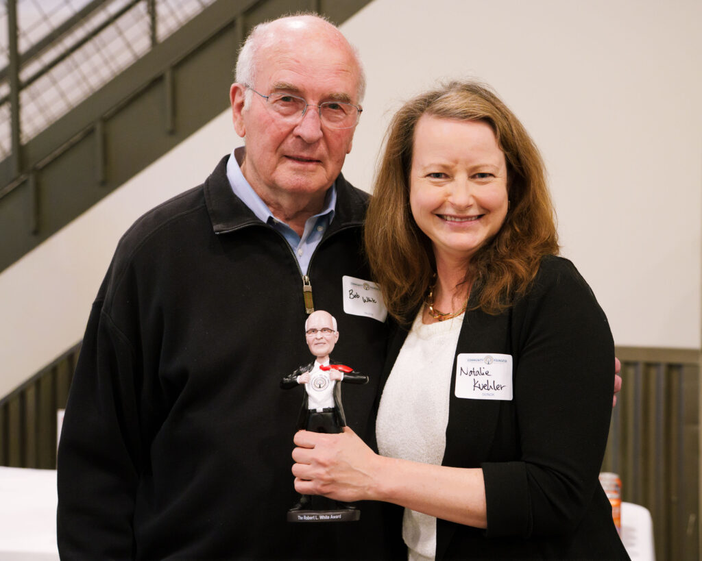 A man and woman smile for a photo at an indoor event with woman holding a small award figurine.